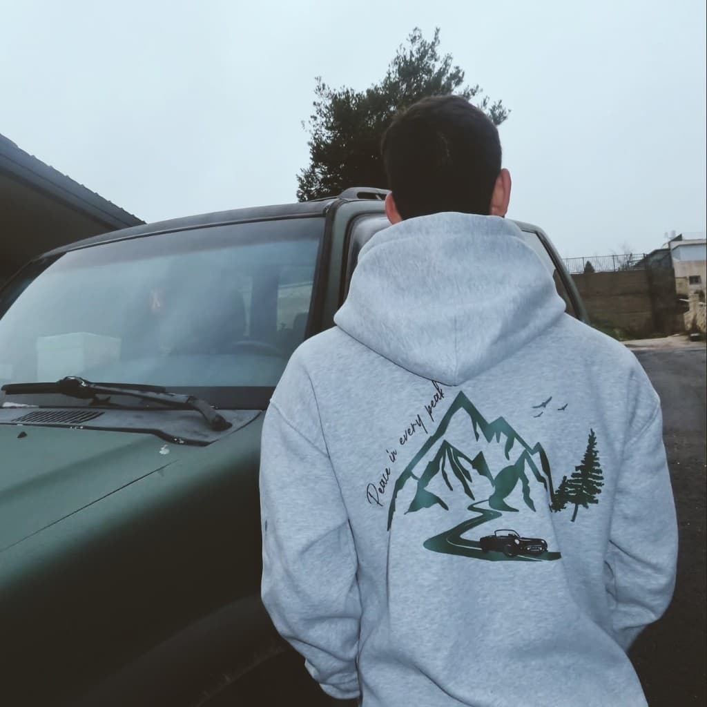 Person from behind in a light grey hoodie with mountain road graphic and Peace in every peak artwork