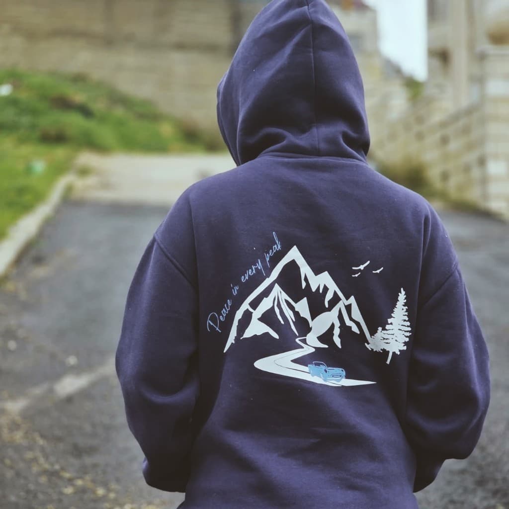 Person from behind in a navy hoodie with hood up; Peace in every peak artwork with mountains, winding road, and car on an uphill street
